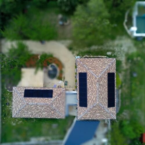 Cette image montre une vue aérienne de deux maisons avec des panneaux solaires installés sur leurs toits. Les bâtiments sont entourés de verdure et d'un aménagement paysager soigné, comprenant des allées et des zones de jardin. Les panneaux solaires sont bien visibles et semblent être installés de manière symétrique sur les toits en tuiles. L'image semble avoir un effet de flou en périphérie, ce qui attire l'attention sur les toits et les panneaux solaires.