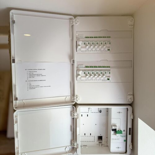 Un tableau électrique Schneider ouvert, installé sur un mur intérieur, affiche plusieurs disjoncteurs organisés en rangées. Deux portes transparentes sont ouvertes, révélant une liste détaillée des circuits électriques. Une partie du tableau semble en attente de composants supplémentaires. L’installation est propre et bien agencée, avec un interrupteur mural visible en dessous.