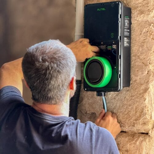 Installation d'une borne de recharge IRVE à Alixan par QR ENERGIES. Un technicien, vêtu d’un t-shirt noir, installe ou effectue la maintenance d’une borne de recharge Autel fixée sur un mur en pierre. Il manipule les composants internes de l’appareil tout en connectant un câble d’alimentation. La borne affiche un anneau lumineux vert, indiquant qu’elle est sous tension ou en fonctionnement.