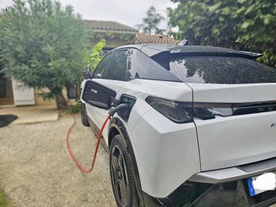 Un SUV électrique blanc est en cours de recharge dans une allée de maison, connecté à une borne via un câble rouge. La carrosserie du véhicule est mouillée, indiquant une récente pluie. En arrière-plan, une maison avec un toit en tuiles et un jardin verdoyant sont visibles, renforçant l’ambiance résidentielle. Le voyant vert sur la prise de recharge confirme que le processus de charge est en cours.