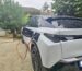 Un SUV électrique blanc est en cours de recharge dans une allée de maison, connecté à une borne via un câble rouge. La carrosserie du véhicule est mouillée, indiquant une récente pluie. En arrière-plan, une maison avec un toit en tuiles et un jardin verdoyant sont visibles, renforçant l’ambiance résidentielle. Le voyant vert sur la prise de recharge confirme que le processus de charge est en cours.