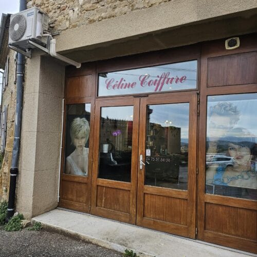 Façade d’un salon de coiffure nommé "Céline Coiffure" dans un bâtiment en pierre. La devanture est en bois avec de grandes vitrines décorées d’images de modèles coiffés. Un climatiseur est fixé sur le mur extérieur, et une plaque avec le numéro 2 est visible à droite de l’entrée. L’intérieur du salon est partiellement visible à travers les vitres, laissant entrevoir un espace aménagé avec des miroirs et du mobilier de coiffure. L’environnement extérieur montre une rue en pente avec quelques bâtiments anciens et un ciel nuageux.