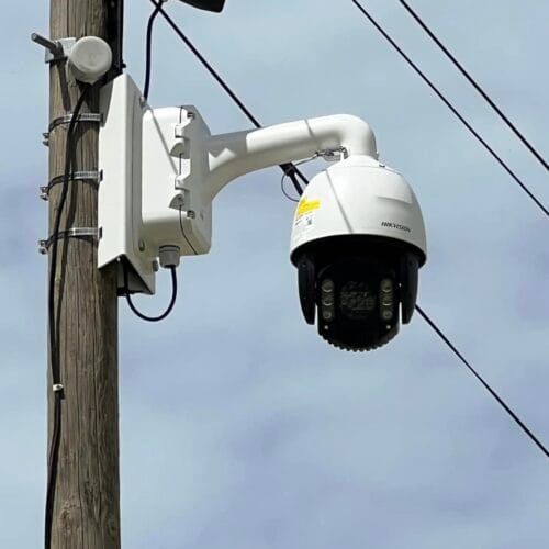 Une caméra de surveillance Hikvision est installée sur un poteau en bois, fixée à un support métallique. Il s'agit d'une caméra motorisée avec vision nocturne, équipée de LED infrarouges pour une surveillance efficace de jour comme de nuit. Des câbles électriques et un boîtier de connexion sont visibles à proximité, reliant l’installation au réseau. En arrière-plan, un ciel légèrement nuageux et des fils électriques traversent l’image.