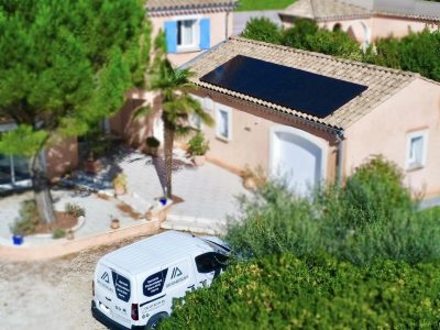 Vue aérienne d’une maison de style méditerranéen avec des murs couleur sable et des volets bleus. Un panneau solaire noir est installé sur le toit du garage. Un véhicule utilitaire blanc, portant le logo "QR Énergies" et des inscriptions indiquant des services en électricité et photovoltaïque, est stationné devant la maison. La propriété est entourée d’arbres, dont un palmier et des arbustes verts, créant un cadre naturel et ensoleillé.
