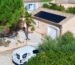 Vue aérienne d’une maison de style méditerranéen avec des murs couleur sable et des volets bleus. Un panneau solaire noir est installé sur le toit du garage. Un véhicule utilitaire blanc, portant le logo "QR Énergies" et des inscriptions indiquant des services en électricité et photovoltaïque, est stationné devant la maison. La propriété est entourée d’arbres, dont un palmier et des arbustes verts, créant un cadre naturel et ensoleillé.