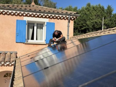 Un installateur travaille sur un toit équipé de panneaux solaires, utilisant une visseuse électrique. Il porte un casque antibruit et des vêtements de travail noirs. En arrière-plan, une maison aux murs orangés avec des volets bleus est visible sous un ciel dégagé. Les panneaux photovoltaïques brillent sous le soleil, reflétant l’environnement.
