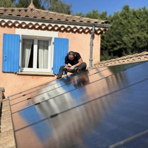 Un installateur travaille sur un toit équipé de panneaux solaires, utilisant une visseuse électrique. Il porte un casque antibruit et des vêtements de travail noirs. En arrière-plan, une maison aux murs orangés avec des volets bleus est visible sous un ciel dégagé. Les panneaux photovoltaïques brillent sous le soleil, reflétant l’environnement.