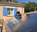 Un installateur travaille sur un toit équipé de panneaux solaires, utilisant une visseuse électrique. Il porte un casque antibruit et des vêtements de travail noirs. En arrière-plan, une maison aux murs orangés avec des volets bleus est visible sous un ciel dégagé. Les panneaux photovoltaïques brillent sous le soleil, reflétant l’environnement.