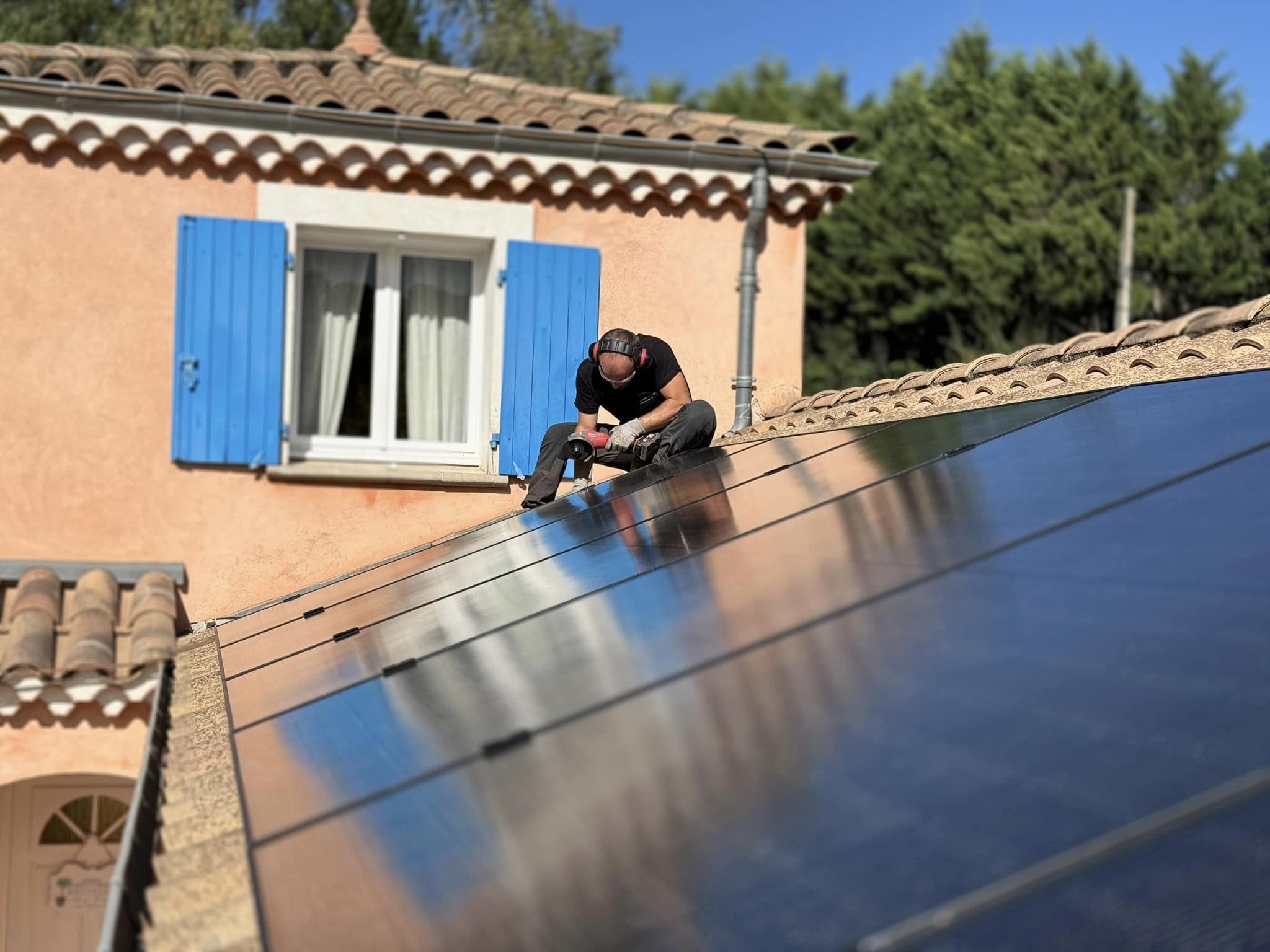 Un installateur travaille sur un toit équipé de panneaux solaires, utilisant une visseuse électrique. Il porte un casque antibruit et des vêtements de travail noirs. En arrière-plan, une maison aux murs orangés avec des volets bleus est visible sous un ciel dégagé. Les panneaux photovoltaïques brillent sous le soleil, reflétant l’environnement.