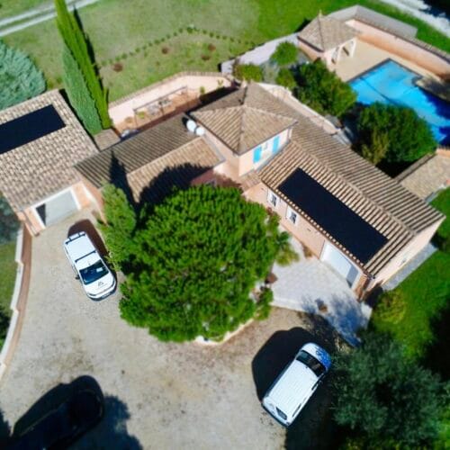 Vue aérienne d’une maison de style méditerranéen avec des murs couleur sable et des volets bleus. Un panneau solaire noir est installé sur le toit du garage. Un véhicule utilitaire blanc, portant le logo "QR Énergies" et des inscriptions indiquant des services en électricité et photovoltaïque, est stationné devant la maison. La propriété est entourée d’arbres, dont un palmier et des arbustes verts, créant un cadre naturel et ensoleillé.
