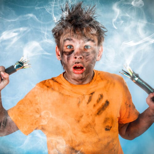 Un jeune homme aux cheveux ébouriffés, le visage couvert de suie, tient deux câbles électriques dénudés fumants dans chaque main. Il porte un t-shirt orange sale avec des traces de main et arbore une expression de surprise mêlée à la peur. La scène évoque un accident électrique, avec une ambiance dramatique renforcée par la fumée et l’arrière-plan bleu.