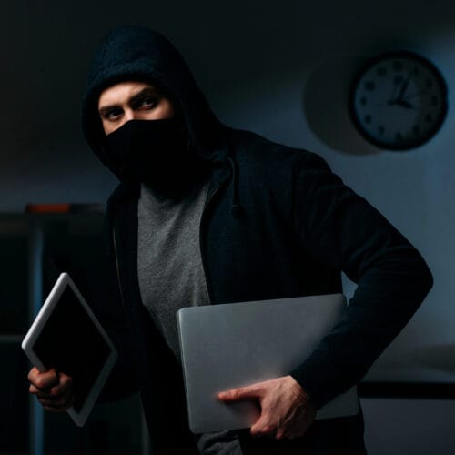 Un voleur vêtu d’un sweat à capuche noir et d’un masque sombre tient un ordinateur portable et une tablette dans un environnement faiblement éclairé. L’ambiance suggère une scène de cambriolage, avec un décor intérieur en arrière-plan comprenant une horloge murale et des étagères légèrement éclairées.