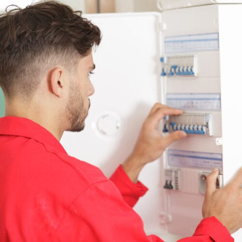 Un électricien en tenue de travail rouge intervient sur un tableau électrique. Il manipule les disjoncteurs avec précision, assurant la maintenance ou l’installation du système. Le coffret électrique blanc est ouvert, révélant plusieurs rangées de disjoncteurs et d’interrupteurs différentiels. L’environnement semble propre et organisé.