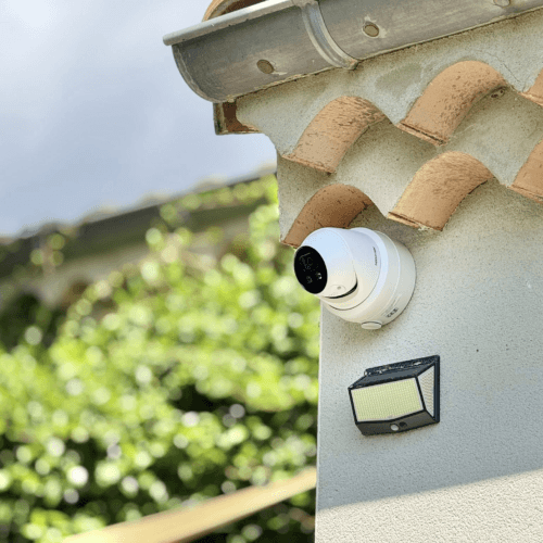 Installation d'alarme à domicile par QR ENERGIES. L’image montre un angle de maison avec une caméra de vidéosurveillance blanche fixée sous les tuiles de rive, orientée vers l’extérieur. Juste en dessous se trouve un projecteur LED rectangulaire avec détecteur de mouvement intégré. L’ensemble est installé sur un mur crépi, probablement pour surveiller l’entrée ou le jardin. À l’arrière-plan, on distingue des tuiles, de la végétation et un ciel partiellement nuageux.
