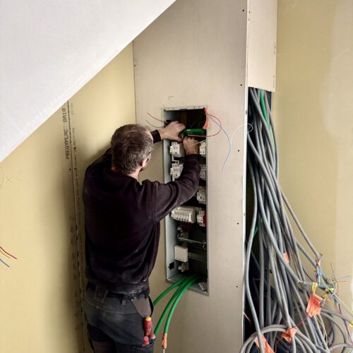 Antoine de la société QR ENERGIES, électricien qualifié et expert en pose de tableaux électriques, en train de passer les câbles et les gaines électriques dans le nouveau tableau électrique installé dans une maison en rénovation à Montélimar. On voit de nombreuses gaines en attente, des gaines télécom, des gaines d'électricité et des murs en placo brut.