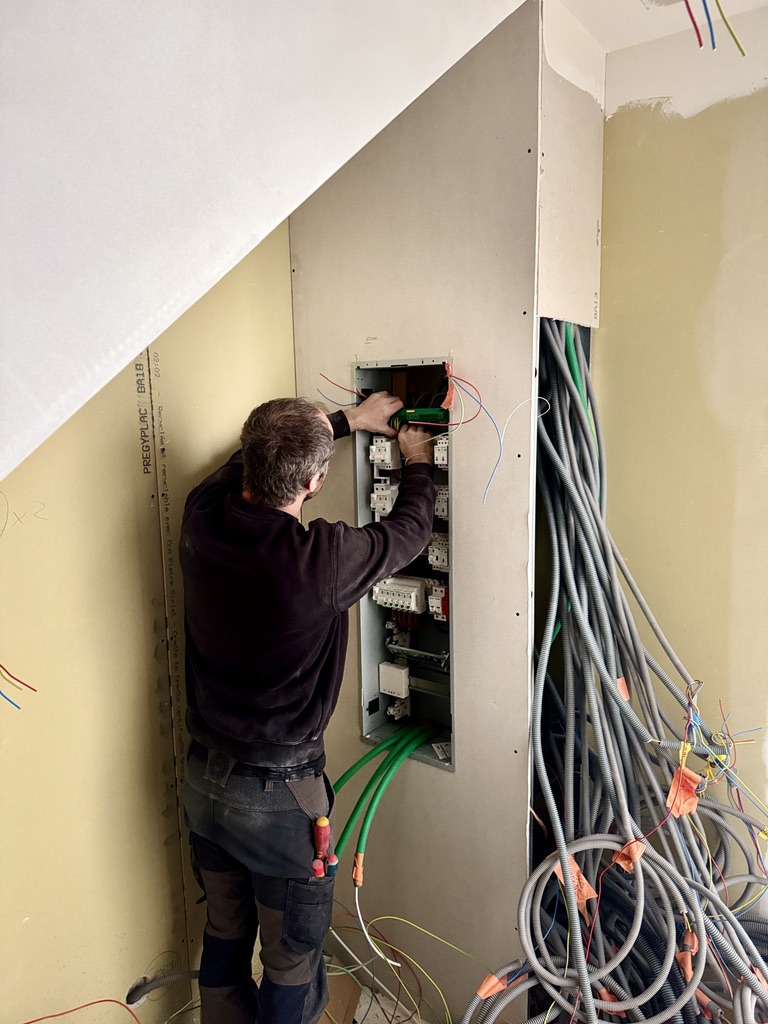Antoine de la société QR ENERGIES, électricien qualifié et expert en pose de tableaux électriques, en train de passer les câbles et les gaines électriques dans le nouveau tableau électrique installé dans une maison en rénovation à Montélimar. On voit de nombreuses gaines en attente, des gaines télécom, des gaines d'électricité et des murs en placo brut.
